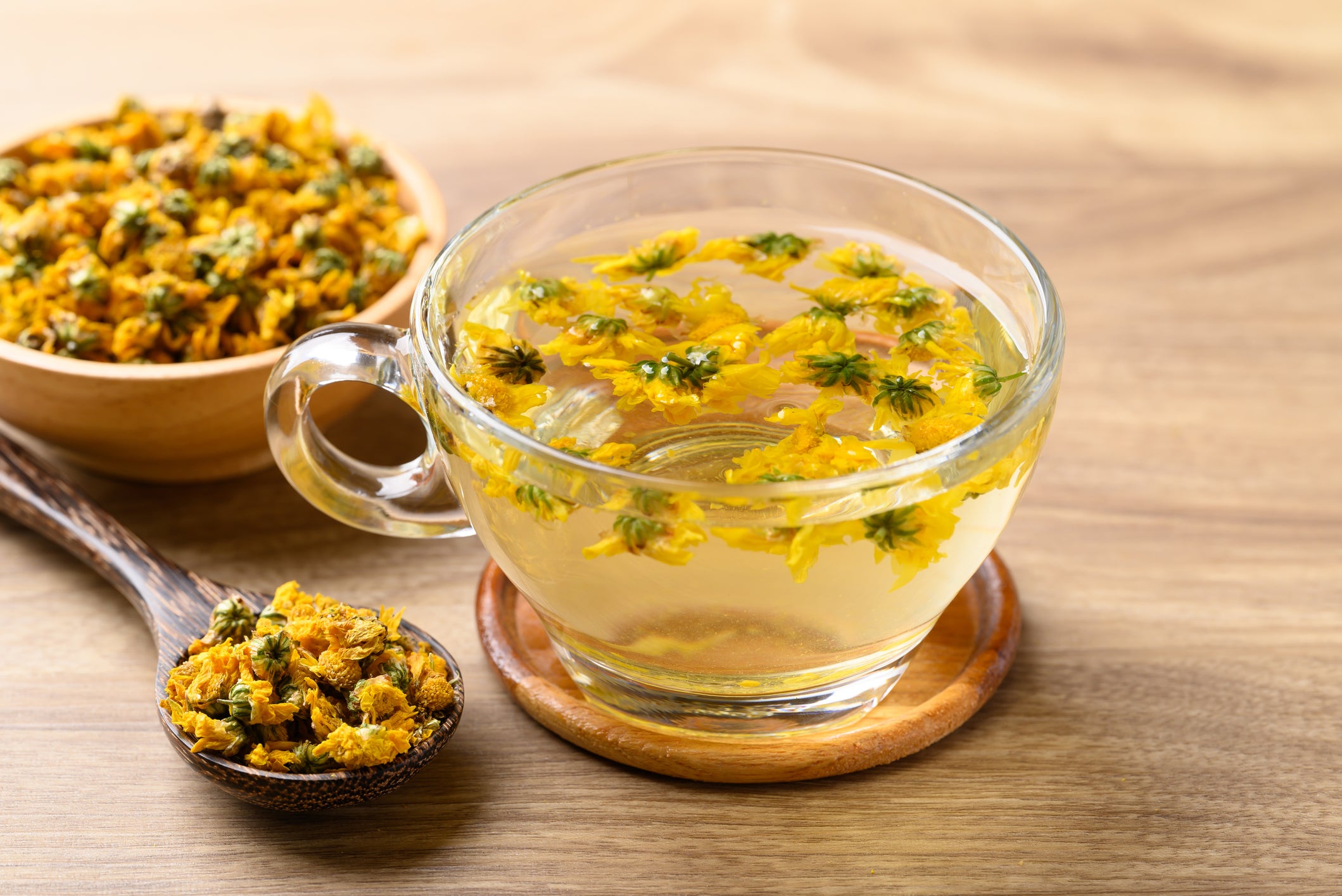 Clear glass cup filled with yellow flower tea on a wooden surface, with a bowl of dried flowers and a spoon nearby.