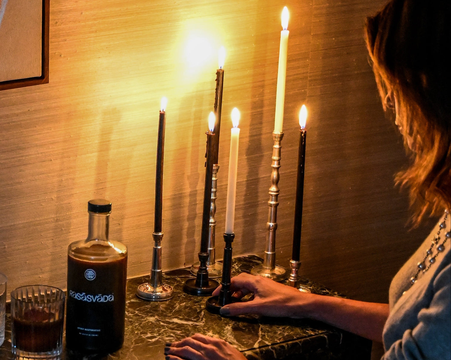 Person lighting candles on a bar counter with a bottle of Rasasvada and glassware.
