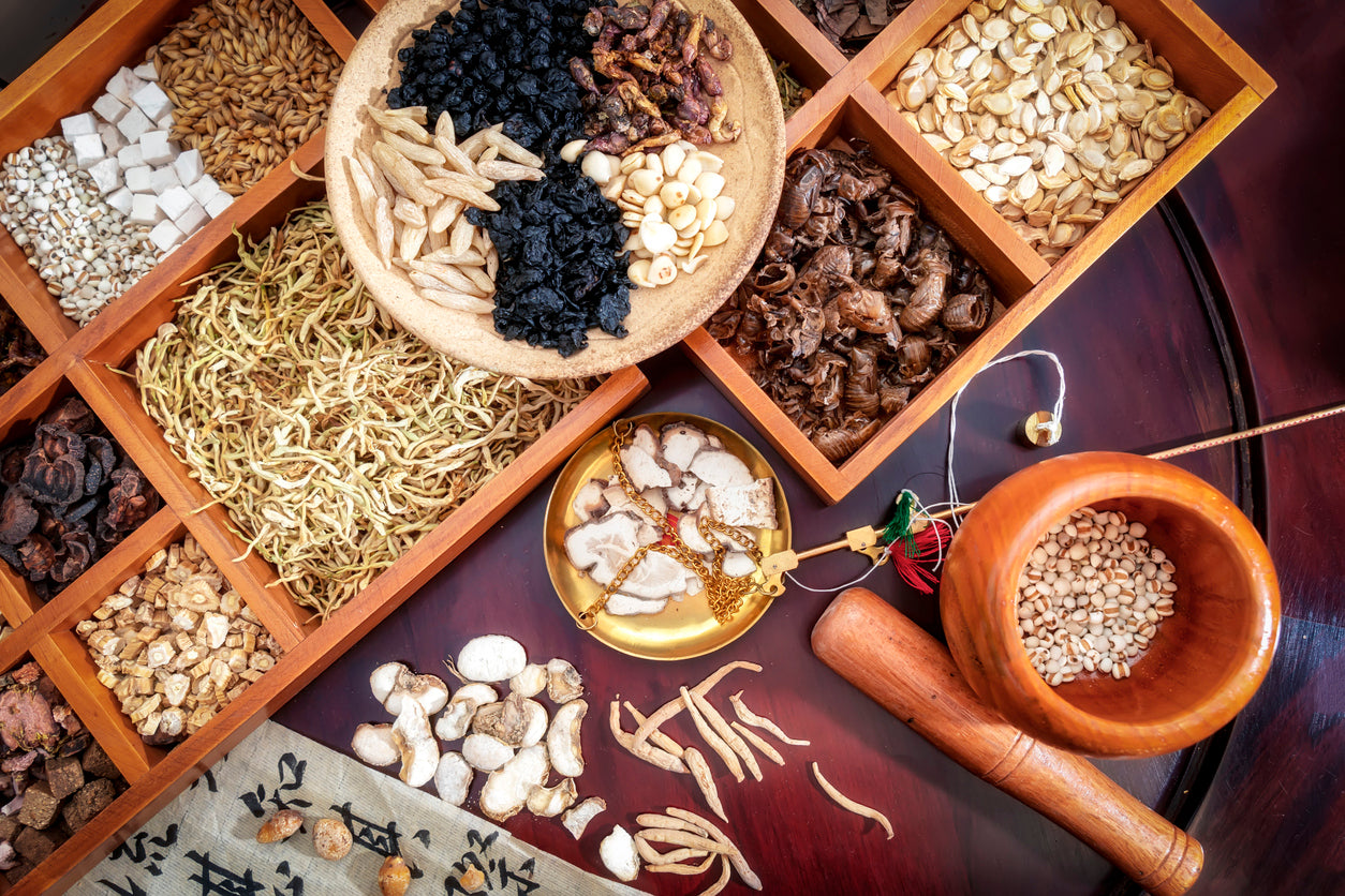 An overhead flat-lay of assorted dried herbal ingredients arranged in a wooden compartment tray, with a small wooden bowl, mortar, pestle, and loose roots on a dark wooden surface.