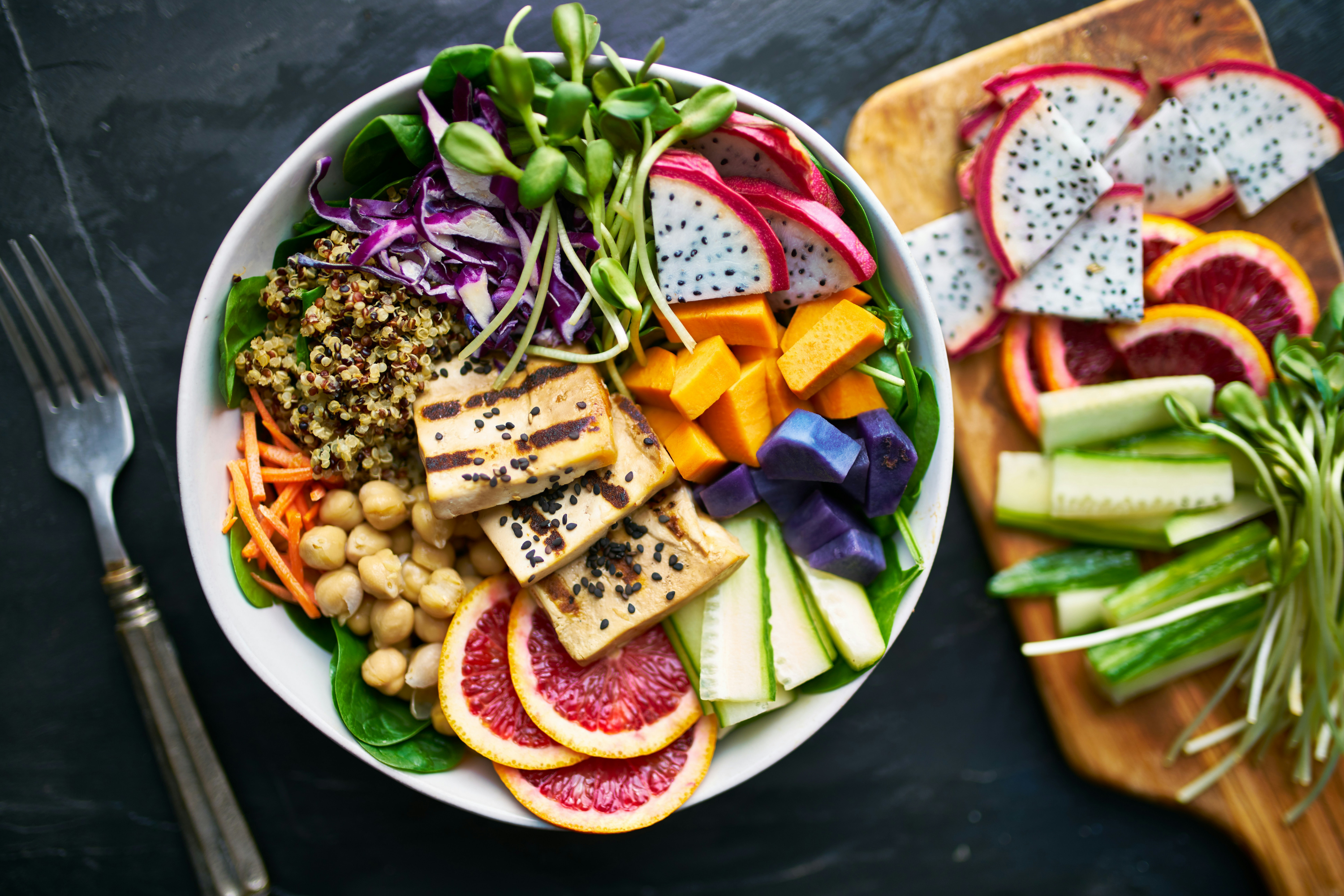 A large white bowl filled with a colorful, healthy salad featuring grilled tofu, chickpeas, quinoa, greens, red cabbage, cucumber, mango, and sliced fruits, presented on a dark slate background with a fork and cutting board.