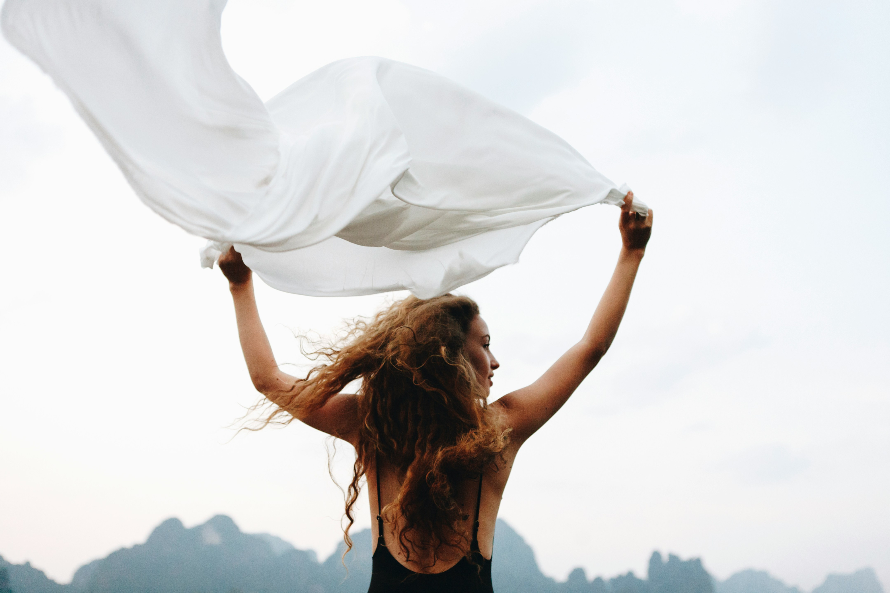 A woman outdoors holding and swirling a large flowing white dress or fabric against a bright sky, with her hair also blowing in the wind.